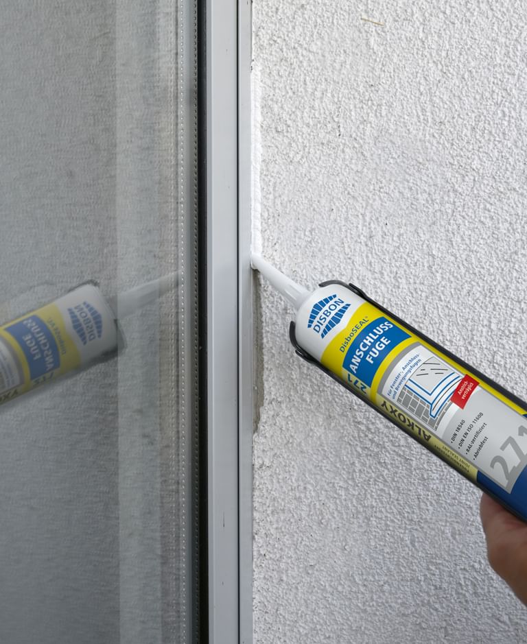 Eine Dichtmasse wird mit einer Kartuschenpistole auf eine Fensterrahmennaht aufgetragen. Die Wand ist hell und strukturiert, und die Dichtmasse wird gleichmäßig entlang der Kante aufgetragen. 